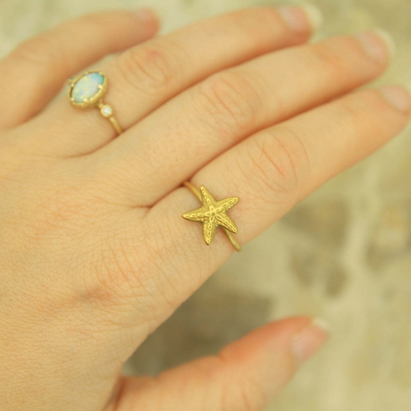 A close-up of a hand wearing a textured yellow gold starfish ring on one finger, with another ring visible on the middle finger, set against a soft, blurred sandy background.
