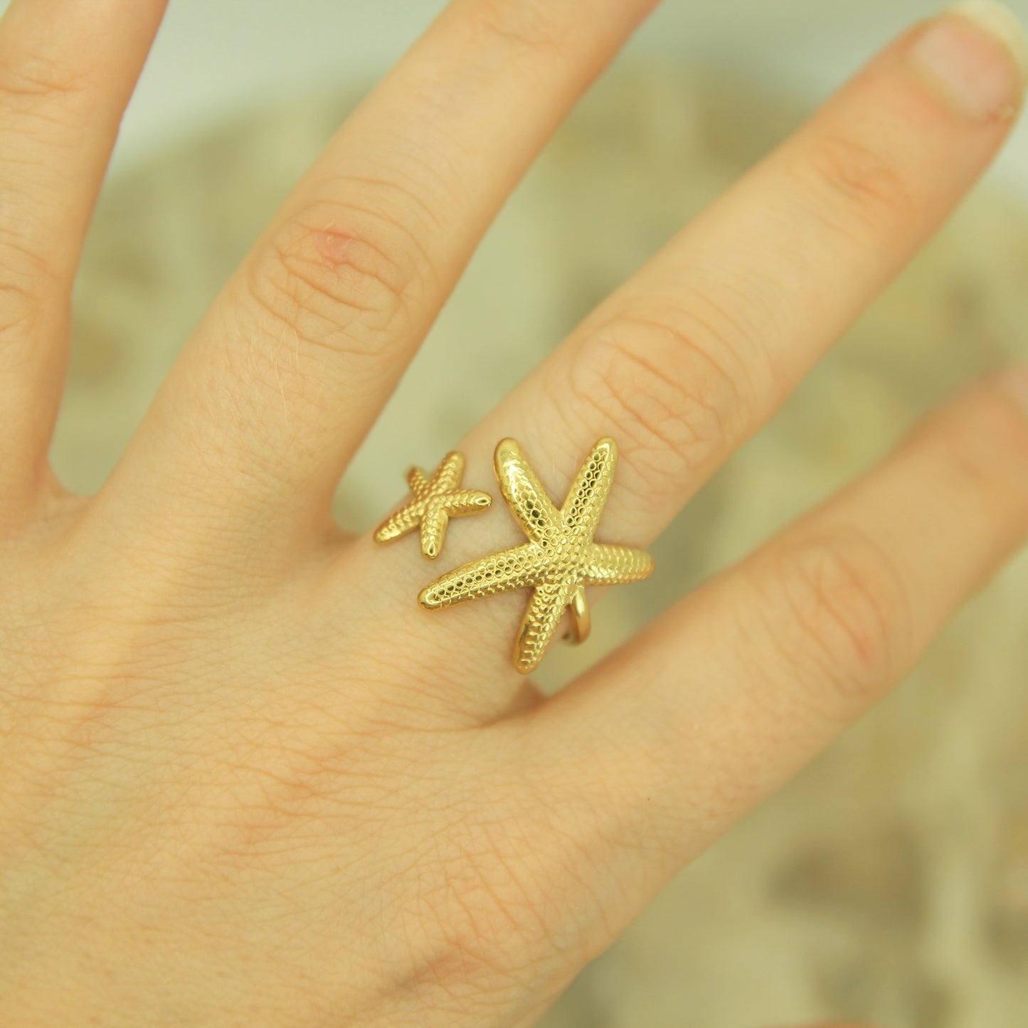 A gold ring featuring two textured starfish, worn on a person's finger against a soft, blurred background.