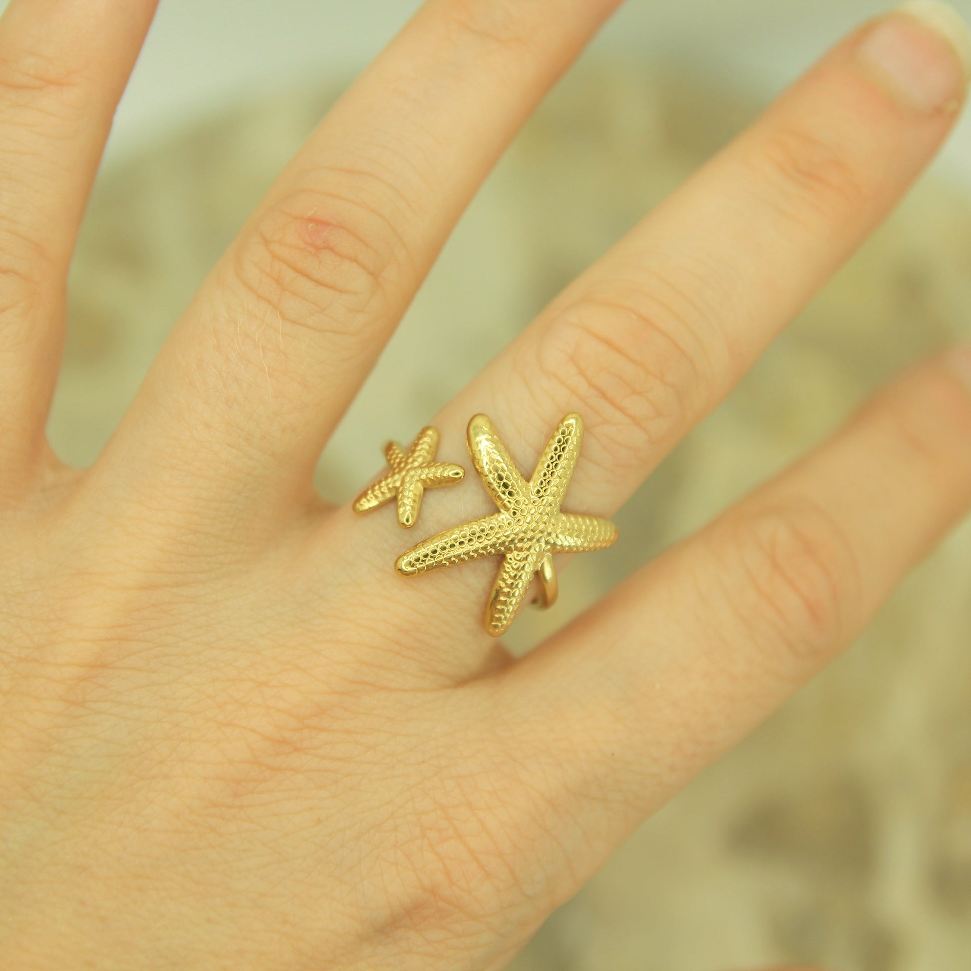 A gold ring featuring two textured starfish, worn on a person's finger against a soft, blurred background.