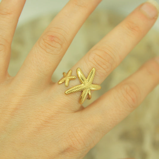 A gold ring featuring two textured starfish, worn on a person's finger against a soft, blurred background.